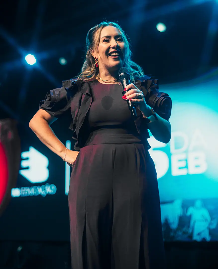 Euza Bispo sorrindo no palco, com o telão da EB Educação ao fundo.
